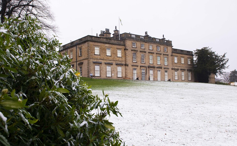 Historic stone building with multiple windows and chimneys, set in a snowy landscape with a green bush dusted in snow in the foreground.